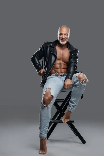 Masculine and stylish male model with striking physique, grey beard, and bare chest, wearing black leather jacket and ripped jeans, sitting on a studio chair against a neutral grey backdrop