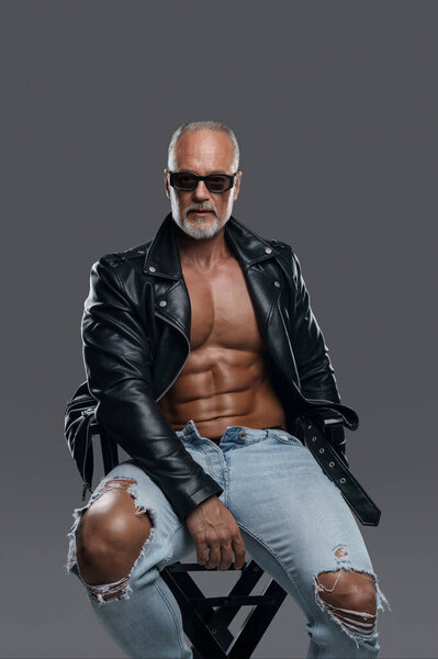 Charismatic muscular male model of mature age with a stylish gray beard and bare torso wearing black leather jacket and ripped jeans, sporting sunglasses while seated on a studio chair
