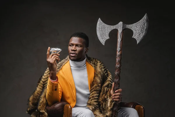 A stylish dark-skinned man in a yellow jacket holding a two-handed axe and a large diamond while sinnting against a dark backdrop