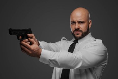 Beyaz gömlekli ve kravatlı kel bir adam, gri bir stüdyo arkaplanına karşı bir dedektif ya da koruma titreşimleri yayarak tabancayı hedef alıyor.