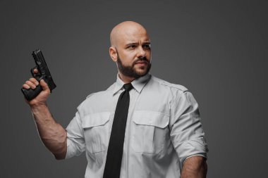 Beyaz tişörtlü ve kravatlı kel bir adam tabanca tutuyor. Gri bir stüdyo geçmişine karşı bir dedektif, koruma ya da güvenlik uzmanının örneğini veriyor.