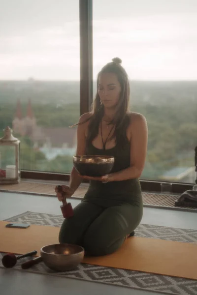 Woman in sportswear practicing spiritual activities in a peaceful environment, meditating with a singing Tibetan bowl in an apartment overlooking a panoramic city skyline