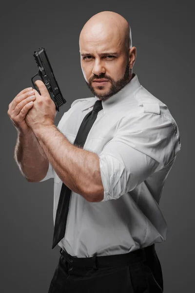 身穿白衬衫 打着领带的秃头男人拿着一支手枪 象征着侦探 保镖或安保专业人员在灰色工作室背景下的形象 图库图片