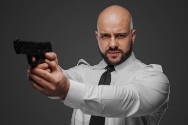 Beyaz gömlekli ve kravatlı kel bir adam, gri bir stüdyo arkaplanına karşı bir dedektif ya da koruma titreşimleri yayarak tabancayı hedef alıyor.
