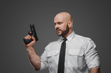 Beyaz tişörtlü ve kravatlı kel bir adam tabanca tutuyor. Gri bir stüdyo geçmişine karşı bir dedektif, koruma ya da güvenlik uzmanının örneğini veriyor.