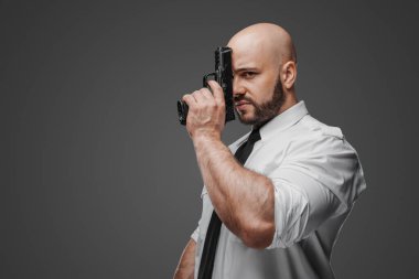 Kel, sakallı, beyaz gömlekli ve kravatlı bir adamın dramatik portresi yüzüne silah dayıyor. Gri bir zemine karşı dedektif, koruma ya da güvenlik temalarını sembolize ediyor.