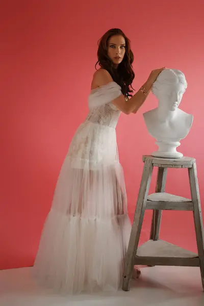 漂亮的黑发姑娘 穿着精致的白色连衣裙 穿着粉色背景的古希腊半身像旁边摆姿势 — 图库照片