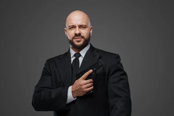 Bald and bearded man in a black suit holds a pistol, exuding power against a gray studio backdrop