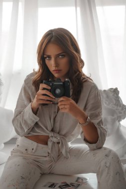 Beyaz bir masa üzerinde antik Yunan büstleri arasında oturan, açık renk elbiseli, şık bir esmer.