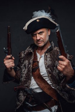 Gri sakallı, kahverengi yelek giymiş, gri bir duvara karşı tek elli iki tüfekle poz veren deneyimli bir korsan.