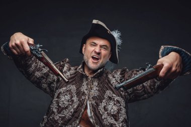 Gri sakallı, kahverengi yelek giymiş, gri bir duvara karşı tek elli iki tüfekle poz veren deneyimli bir korsan.