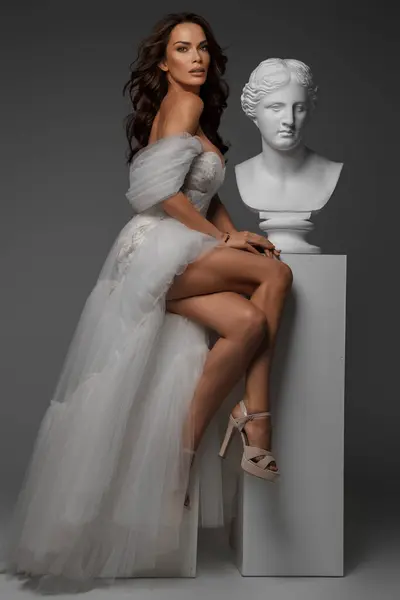 身着白色长袍的优雅女人与古典希腊雕像并排摆放在摄影棚中 — 图库照片