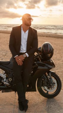 Siyah takım elbiseli şık bir adam bulutlu bir günbatımına karşı ıssız bir plajda spor motosikletine yaslanıyor.