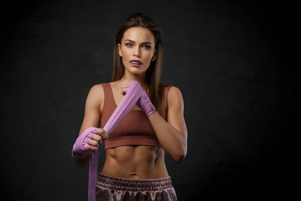 Stunning woman in sports bra and shorts, with boxing wraps and with mouthguard against a dark wall
