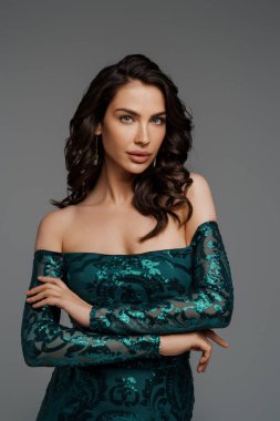 Zengin yeşil gece elbisesi içinde zarif esmer manken. Gri bir arka planda gür saç stili sergiliyor.