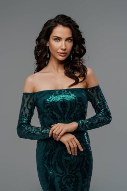 Zarif yeşil bir gece elbisesi içinde, gri bir arka planda, şehvetli bir saç stiliyle göz kamaştırıcı bir esmer.
