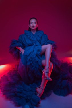 Lüks mavi bir karnaval elbisesi içindeki güzel manken, neon ışıklarıyla vurgulanan sıcak ışıklı bir stüdyoda poz veriyor.