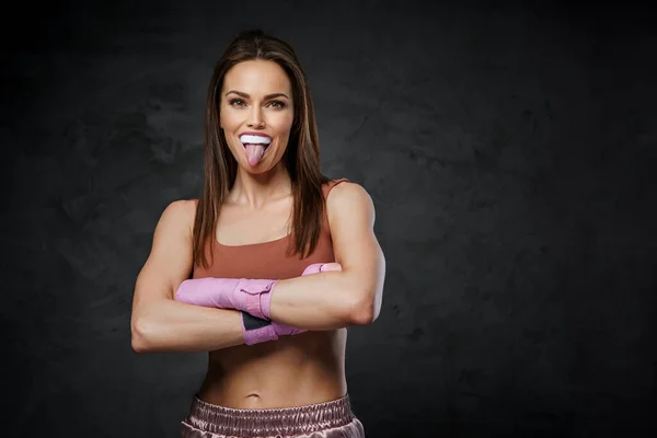 Cheerful woman in sports bra and shorts, biting a boxing mouthguard, with hand wraps against a dark wall