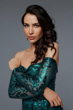 Zarif yeşil bir gece elbisesi içinde, gri bir arka planda, şehvetli bir saç stiliyle göz kamaştırıcı bir esmer.