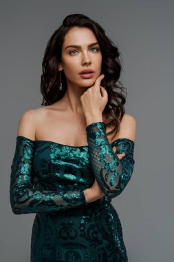 Zengin yeşil gece elbisesi içinde zarif esmer manken. Gri bir arka planda gür saç stili sergiliyor.