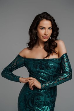 Zengin yeşil gece elbisesi içinde zarif esmer manken. Gri bir arka planda gür saç stili sergiliyor.