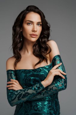 Zengin yeşil gece elbisesi içinde zarif esmer manken. Gri bir arka planda gür saç stili sergiliyor.