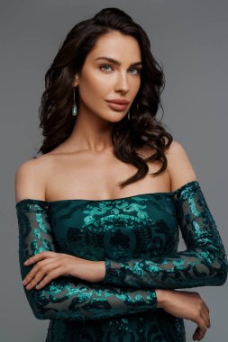 Zarif yeşil bir gece elbisesi içinde, gri bir arka planda, şehvetli bir saç stiliyle göz kamaştırıcı bir esmer.