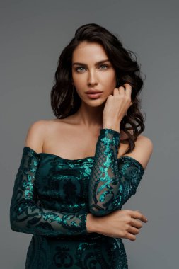 Zarif yeşil bir gece elbisesi içinde, gri bir arka planda, şehvetli bir saç stiliyle göz kamaştırıcı bir esmer.