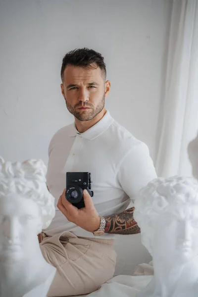 Wealthy man in a white shirt sporting a tattoo, poses with a camera among ancient Greek sculptures in a softly lit room