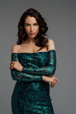 Zarif yeşil bir gece elbisesi içinde, gri bir arka planda, şehvetli bir saç stiliyle göz kamaştırıcı bir esmer.