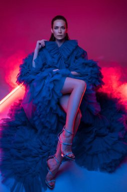 Lüks mavi bir karnaval elbisesi içindeki güzel manken, neon ışıklarıyla vurgulanan sıcak ışıklı bir stüdyoda poz veriyor.