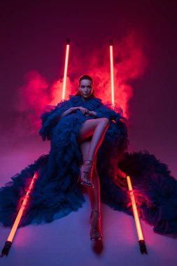 Gösterişli mavi bir karnaval elbisesiyle stüdyoda neon ışıklarının sıcaklığıyla zarif poz veren çarpıcı bir manken..