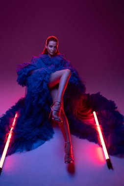 Model görünümlü zarif bir kadın model, şehvetli mavi bir karnaval kıyafeti giyer, neon ışıklı stüdyosunun sıcak atmosferini büyüleyici gülümsemesiyle yakalar.
