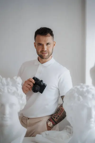 Wealthy man in a white shirt sporting a tattoo, poses with a camera among ancient Greek sculptures in a softly lit room