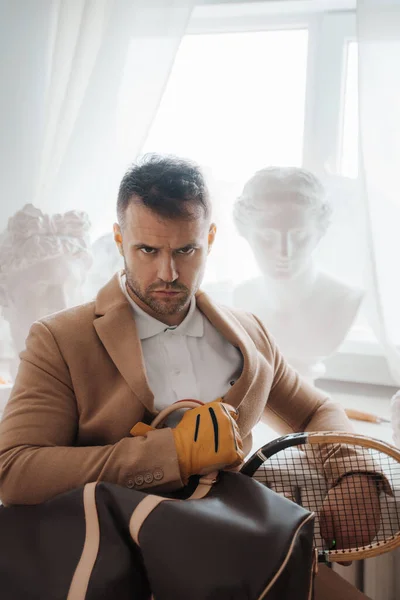 Stylish male model embodies the old-money look, posing with a bag and tennis racket among classical Greek sculptures in a sculptors atelier
