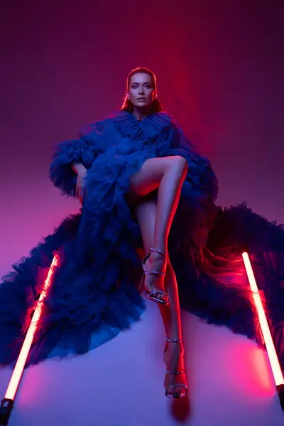 stock image Beautiful model in a lavish blue carnival gown poses in a warmly lit studio, accentuated by neon lights