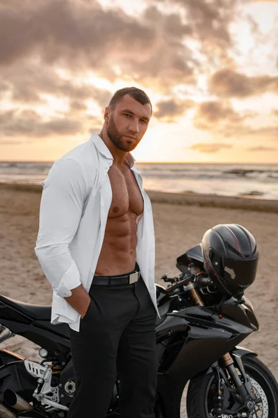 Handsome man, open white shirt revealing toned torso, poses with his black sports bike on a desolate beach, moody sunset sky behind