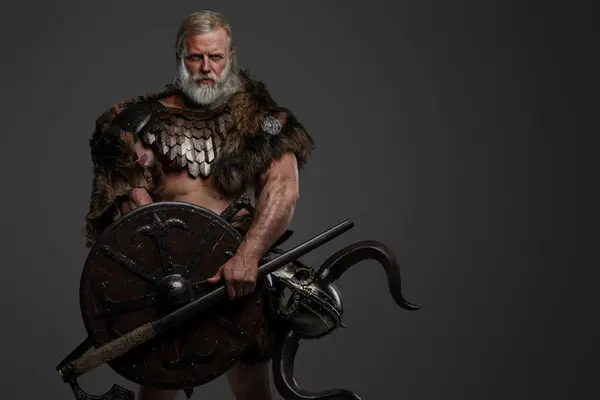 A rugged, silver-haired, bearded elderly viking dressed in fur and lightweight armor, with a helmet attached to his belt, holding an axe and shield against a gray background