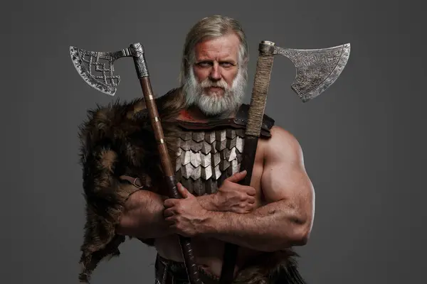 Weathered and venerable Viking hero, donning fur and lightweight armor, sporting a belt-mounted helmet, gripping two axes against a neutral backdrop