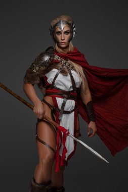 Kırmızı pelerinli, kaslı, sarışın bir Viking kadın, gri arka planda mızrak ve kalkan tutuyor.