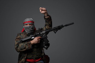Orta Doğulu fanatik bir asker, beyaz keffiyeh giyiyordu ve kamufle olmuş bir üniforma giyiyordu, tüfekle silahlıydı, gri bir arka plana karşı elini kaldırmış poz veriyordu.