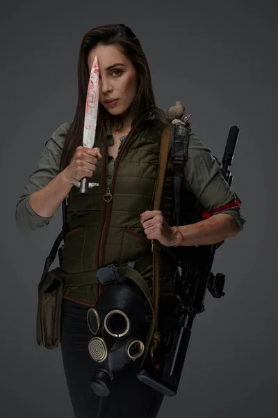 Middle Eastern descent in post-apocalyptic survival gear, brandishing a dagger on a gray backdrop, showcasing her determination to survive