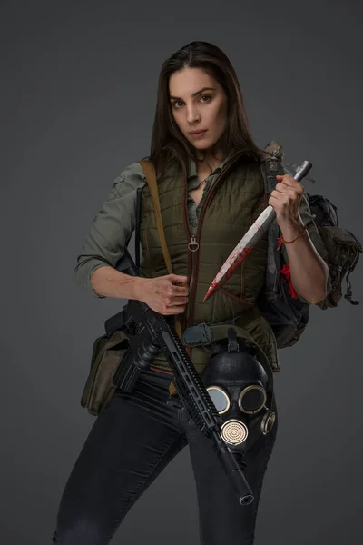 Middle Eastern woman descent in post-apocalyptic survival gear, brandishing a dagger on a gray backdrop, showcasing her determination to survive
