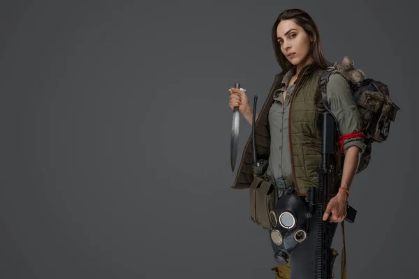 Middle Eastern woman descent in post-apocalyptic survival gear, brandishing a dagger on a gray backdrop, showcasing her determination to survive