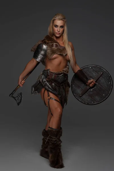 A fierce barbarian or Viking maiden with blonde hair wears lightweight armor, fur, and exposes her chest. She proudly holds an axe and shield against a gray background
