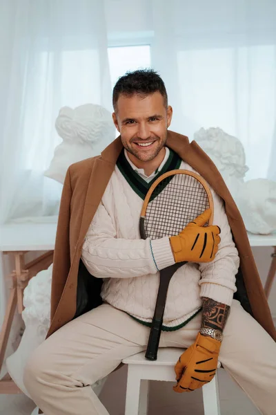 Charming man in old money style, sporting a captivating smile, poses with a tennis racket amid ancient Greek sculptures