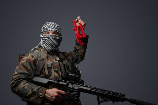 Ближневосточный солдат-фанатик радикалов, одетый в белую кеффию и камуфляжную полевую форму, вооруженный винтовкой, поднял руку, держа красную тряпку и пульт дистанционного управления