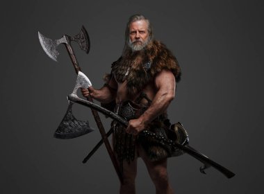 Kürk ve hafif zırh giymiş yaşlı sakallı Viking savaşçısı iki büyük balta tutuyor, gri bir arka plana karşı.