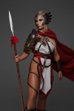 Kırmızı pelerinli, kaslı, sarışın bir Viking kadın, gri arka planda mızrak ve kalkan tutuyor.