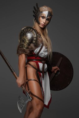 Çıplak bacaklı fantezi zırhlı Viking kadın. Gri bir arka planda kendine güvenen bir savaş baltası ve kalkanı var.